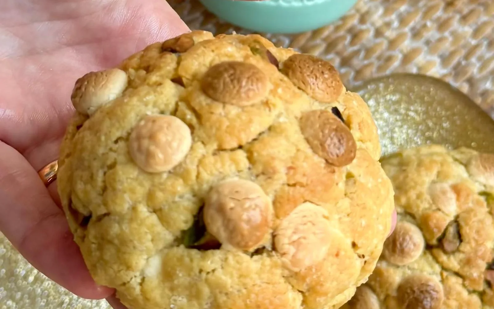 A pistachio heart chunky cookie held in a hand, with a second cookie and a bowl of Lindt pistachio bonbons behind it