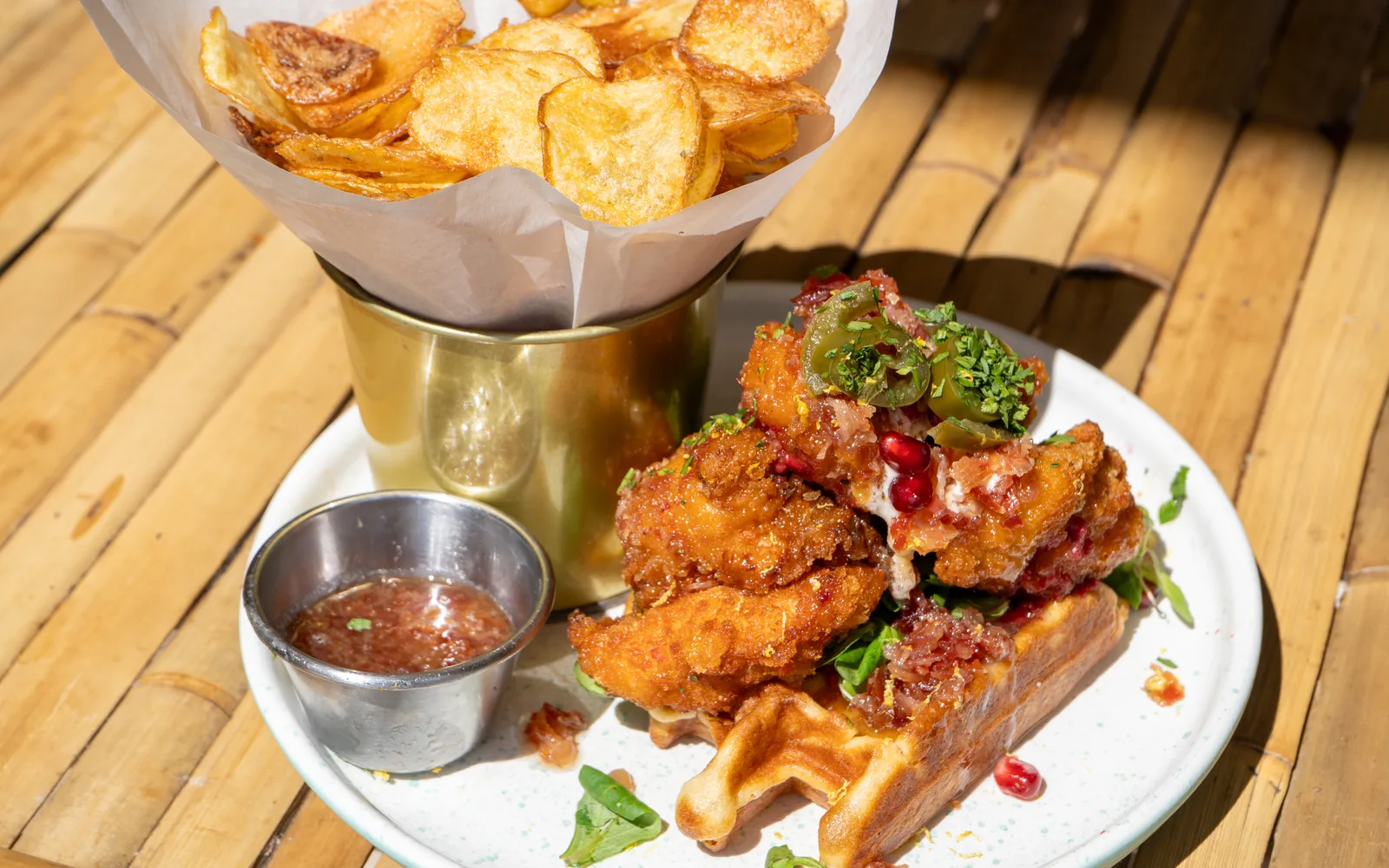 The whole Chicken & Waffles Stack plate on a table at Cookie Beacon Budapest — the chicken and waffle beside a small pot of sauce, with a paper cone of homemade potato chips in a brass cup