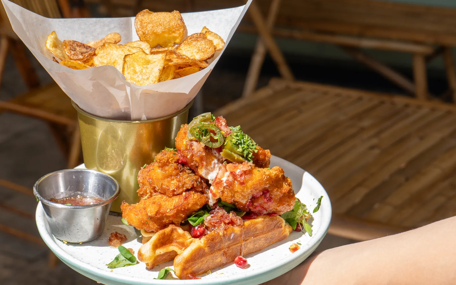 The Chicken & Waffles Stack carried to a terrace table at Cookie Beacon Budapest — panko chicken stacked on a golden waffle with jalapeño and parsley on top, a cone of homemade potato chips and a pot of sauce alongside
