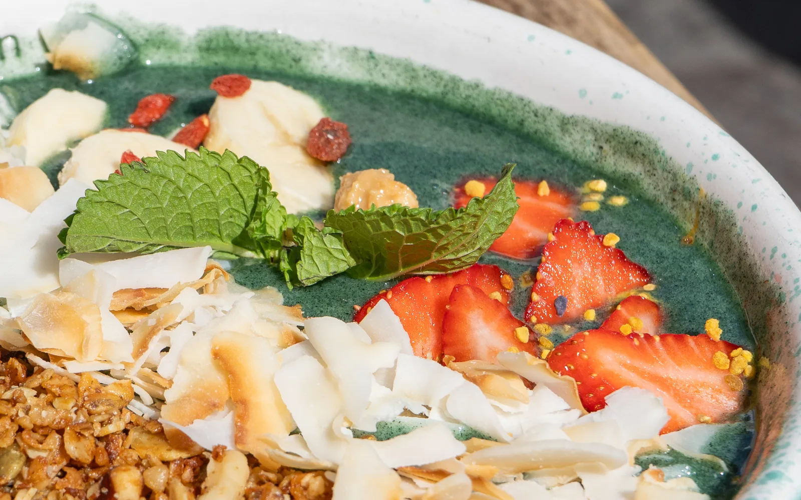 Close-up of the Spirulina Power Granola Bowl at Cookie Beacon Budapest: cut strawberries scattered with yellow pollen granules, a sprig of mint, goji berries and banana pieces on the teal-green spirulina base, with toasted coconut flakes and honey toasted granola at the front