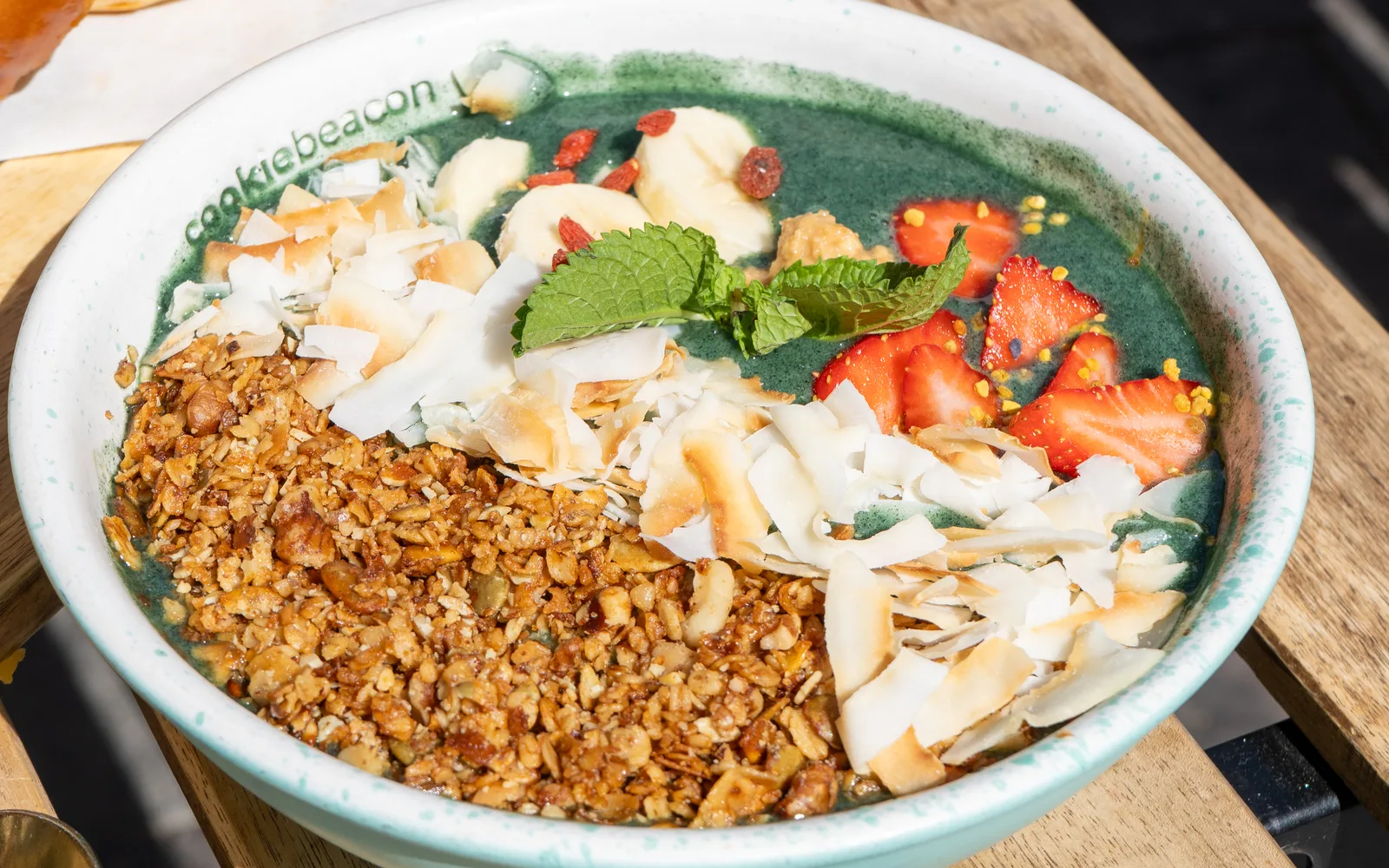 The Spirulina Power Granola Bowl on a wooden terrace table at Cookie Beacon Budapest: a white bowl filled with a teal-green spirulina base, a heap of honey toasted granola, toasted coconut flakes, banana pieces, goji berries, cut strawberries scattered with yellow pollen granules and a sprig of mint