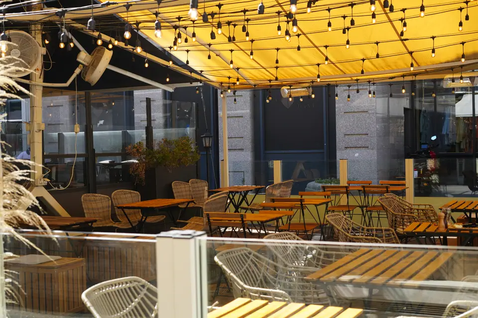 Slatted wooden tables and rattan chairs on the terrace at Cookie Beacon Kralja Petra in Belgrade, under a yellow awning strung with festoon lights