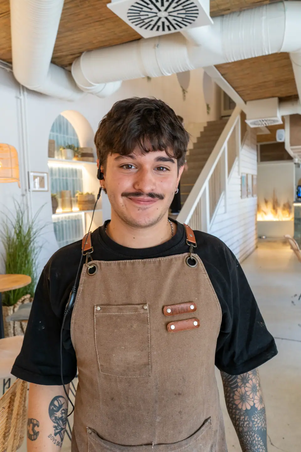 A Cookie Beacon staff member in a brown canvas apron and a headset, standing on the ground floor of the Ferenciek tere café with the lit arched shelving alcove and the staircase behind
