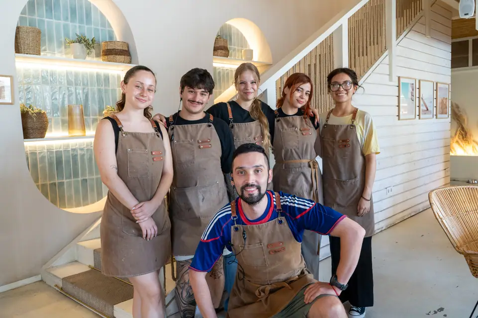 Six Cookie Beacon staff in brown canvas aprons standing together on the ground floor of the Ferenciek tere café, in front of the lit glass-brick alcove and the staircase
